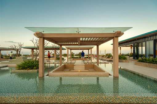 A modern outdoor pool area with a wooden pergola and people walking around.
