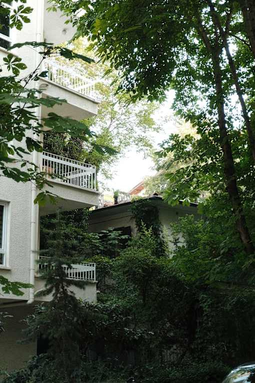 A white building with balconies is surrounded by trees and greenery.
