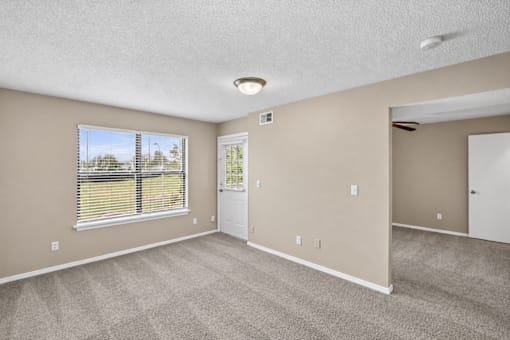 A room with a carpeted floor, a window with blinds, and a door leading to another room.