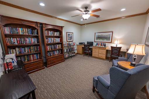 A room with a carpeted floor, a ceiling fan, and a bookshelf filled with books.
