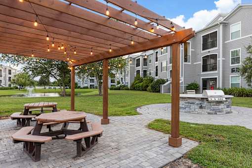A wooden pergola with string lights is over a picnic table in a courtyard.