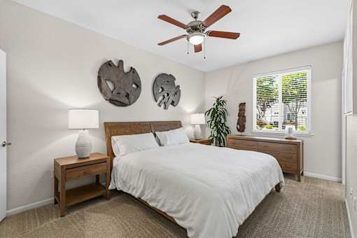 A bedroom with a bed, a nightstand with a lamp, a ceiling fan, and two wall decorations.