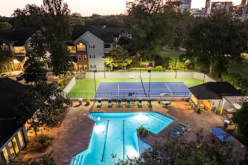 A tennis court is lit up at night with a pool in the foreground.