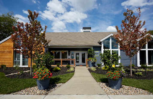 A house with a grey roof and a white door is surrounded by trees and flower pots.