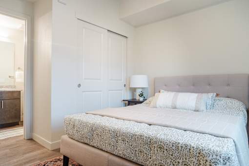 A bed with a white patterned blanket and two white pillows sits in a room with a wooden floor and white walls.
