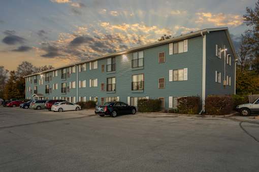 A parking lot in front of a grey apartment building with cars parked.