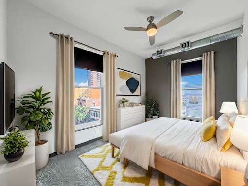 A bedroom with a large bed, a ceiling fan, and a view of a cityscape through the windows.