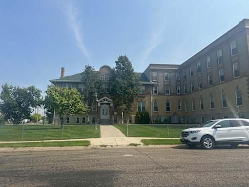 A white car is parked in front of The Cornerstne Apartments with a fence and trees.