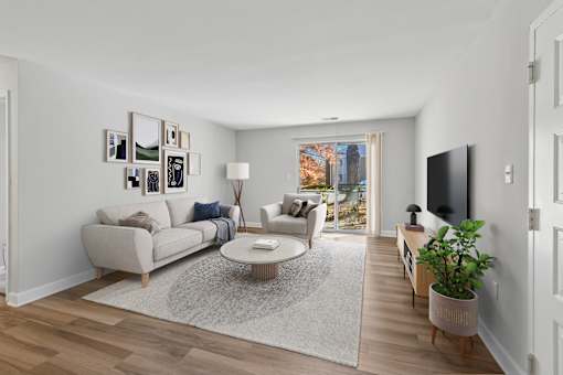 A living room with a grey rug, a white sofa, a grey armchair, a wooden coffee table, a potted plant, and a flat screen TV at Poplar Glen Apartments, Columbia, MD, 21044.