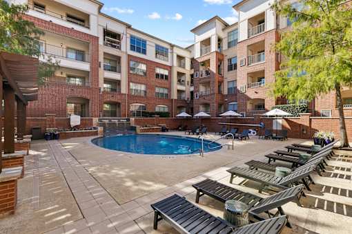 A large outdoor pool area with sun loungers and umbrellas in front of apartment buildings.