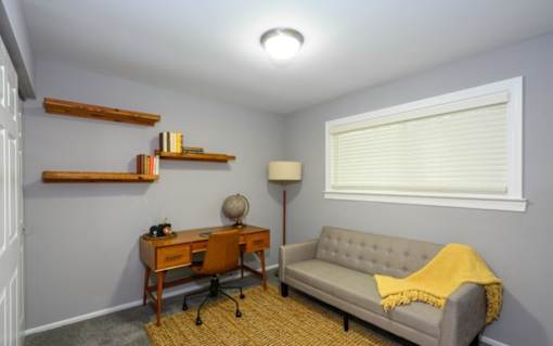 A room with a grey couch, a desk with a chair, a lamp, and a window with blinds.