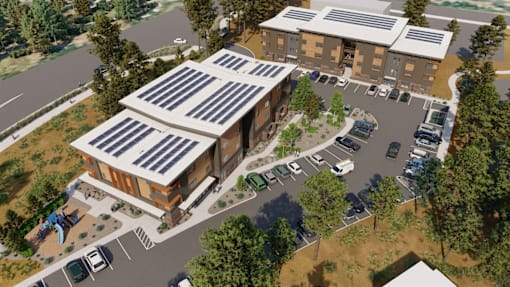 A building with solar panels on the roof is surrounded by a parking lot and trees.