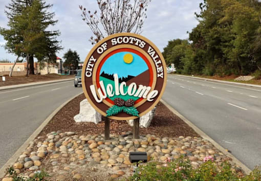 A welcome sign for the City of Scotts Valley is displayed in front of a road.