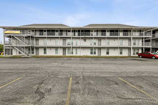 A parking lot in front of a white building with a red car parked in the lot.
