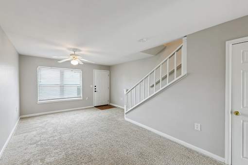A room with a carpeted floor, a ceiling fan, and a staircase.