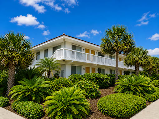 799 - A white building with a balcony is surrounded by green plants.