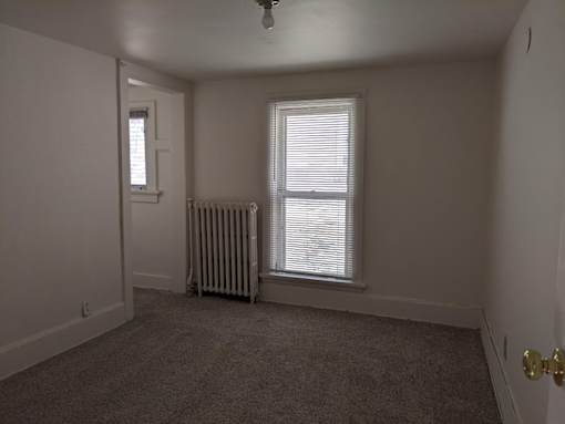 A room with a carpeted floor, a window with blinds, a door, and a radiator.