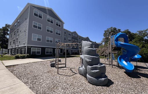 A playground with a blue slide and grey sandbox in front of a grey building.