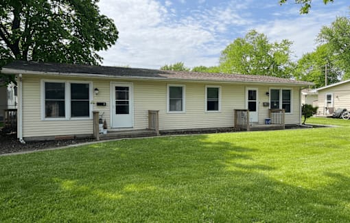 A house with a white siding and a black door is surrounded by a green lawn.