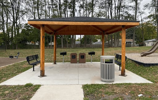 A wooden pavilion with a black roof is surrounded by a concrete walkway and a metal trash can.