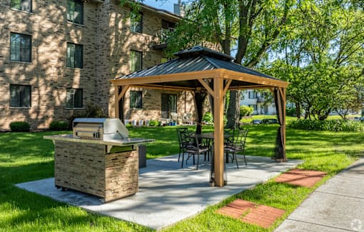 A wooden gazebo with a grill is situated in a grassy area in front of a building.