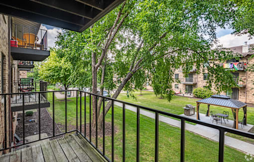 A balcony with a black railing and a wooden floor overlooks a green lawn and trees.