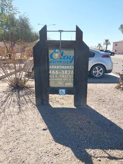 A wooden sign for "Cay Village Apartments" stands in the desert.