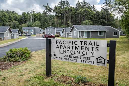 A sign for Pacific Trail Apartments in Lincoln City is displayed in front of a row of houses.