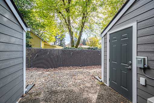 A grey door is open to a backyard with a gravel area and a tree.