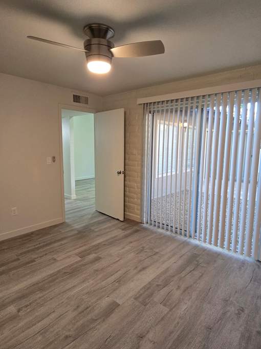 A room with a ceiling fan and light, wooden flooring, and a sliding door with blinds.
