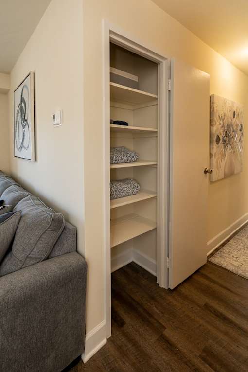 A grey couch sits next to a white wall with a shelf and a painting on it.
