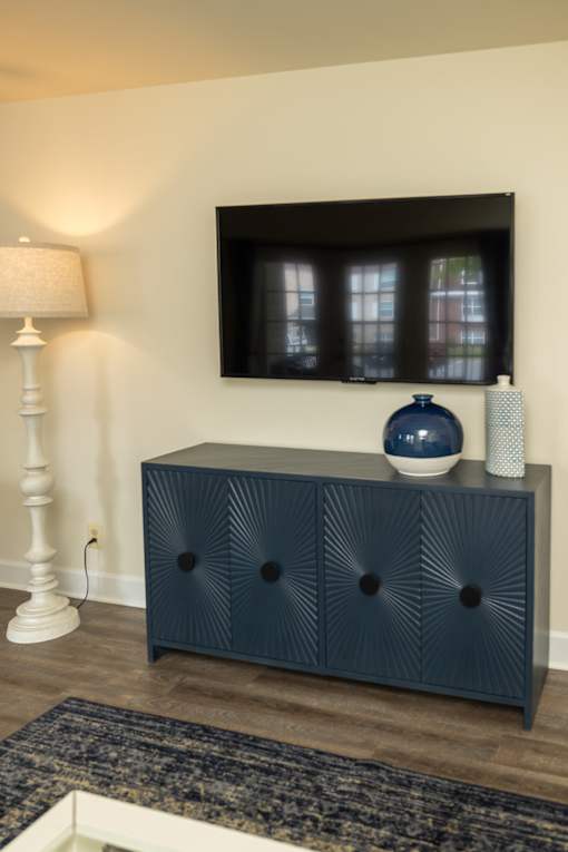 A dark blue cabinet with a vase on top is in front of a wall with a flat screen TV mounted on it at The Brittany Apartments, Pikesville, MD, 21208