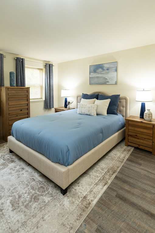 A bed with a blue cover sits in a room with a wooden dresser and a painting on the wall.