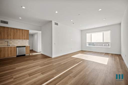 A spacious room with wooden flooring and a kitchen area with white walls.