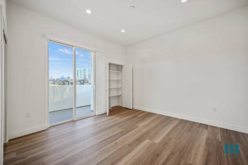 A room with a sliding glass door leading to a balcony, wooden flooring, and a white wall with a shelf.