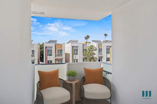 Two chairs with orange pillows are in the foreground of a balcony with a view of apartment buildings and palm trees.