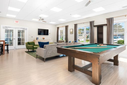 A community lounge with hardwood-style flooring, a comfortable sofa, chairs, a billiards table, a TV, large windows, and two ceiling fans.