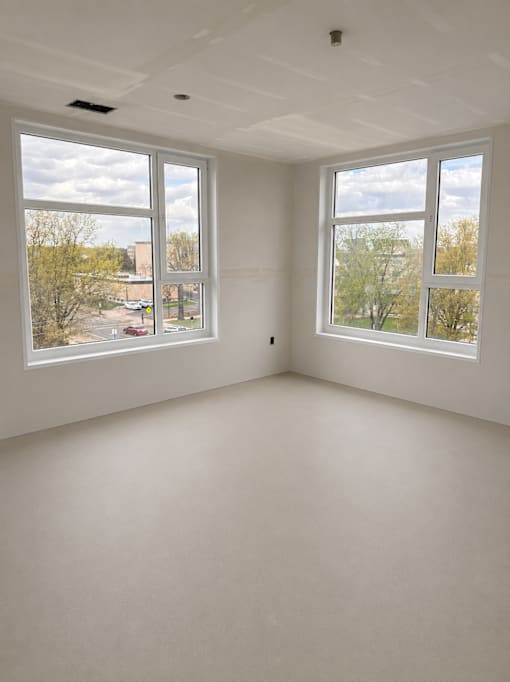 A room with two large windows and a view of trees and a building outside.