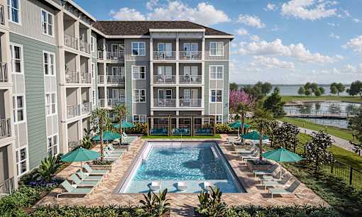 A large swimming pool surrounded by lounge chairs and umbrellas in front of a multi-story apartment building.