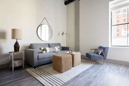 A living room with a grey couch, a round mirror, a lamp, a chair, and a wicker ottoman.