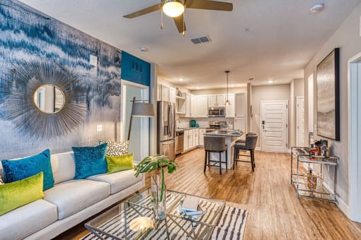 Apartments for Rent in Maitland, FL - The Parker at Maitland Station - Living Room with Wood Vinyl Plank Flooring, Area Rug, Couch, Standing Lamp, Glass-Top Coffee Table, Ceiling Fan, and View of Kitchen.