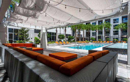 Pet-Friendly Luxury Apartments in Orlando, FL - NORA - An Outdoor Lounge Featuring Dark Wicker Couches, Ottomans, Orange Cushions, Canvas Canopy, String Lights, and View of the Pool Area.