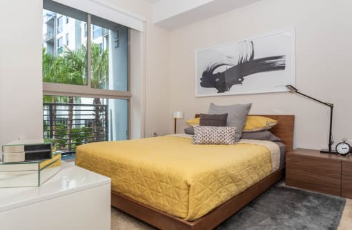One-Bedroom Luxury Apartments in Orlando, FL - NORA - Bedroom with Carpeted Flooring, Area Rug, Large Bed, Nightstand with Lamp, White Dresser, and Expansive Window.