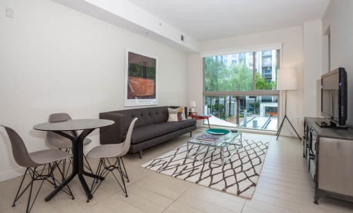 Two-Bedroom Luxury Apartments in Orlando, FL - NORA - Living Room with Light Wood-Style Flooring, Area Rug, Sofa, End Table with Lamp, Glass Top Coffee Table, Round Table with Chairs, Media Cabinet, TV, Standing Lamp, and Expansive Window.