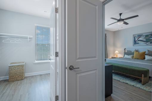 A room with a bed, a wicker basket, a window with blinds, a wall hanging, a ceiling fan, and a door.