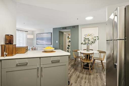 A kitchen with a refrigerator, a dining table with chairs, and a bowl of fruit on the counter.