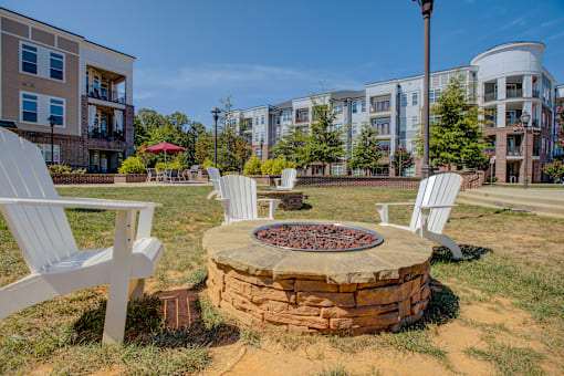 A fire pit sits in the middle of a grassy area with white chairs around it.
