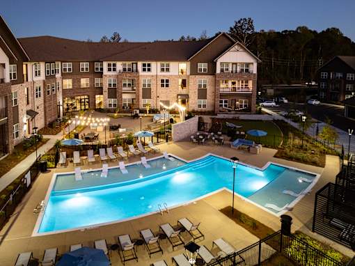 A large swimming pool surrounded by lounge chairs and umbrellas in front of a multi-story apartment building.
