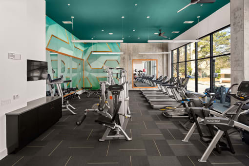 Decatur, GA, Apartments near Glenwood Elementary - Arlo - Fitness Center With Treadmills, Ellipticals, Weight Machines, And Free Weights Along Floor To Ceiling Windows And A Teal Accent Wall.