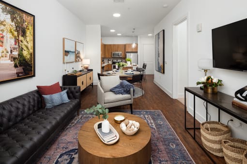 Decatur, GA, Apartments Near Marta Station - Arlo - Living Room With Black Sofa, Round Wood Coffee Table, Wall-Mounted Television, Dining Area, And Modern Kitchen In The Background.